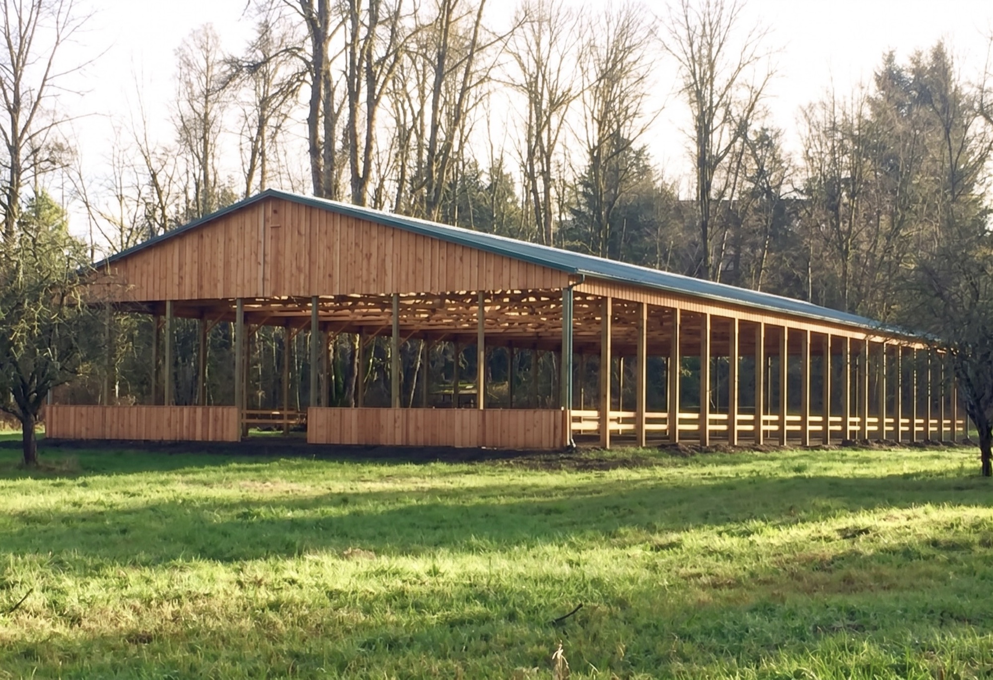 Large cedar-sided open pole building with a green metal roof on a wooded Oregon property, built by Miner Pole Buildings